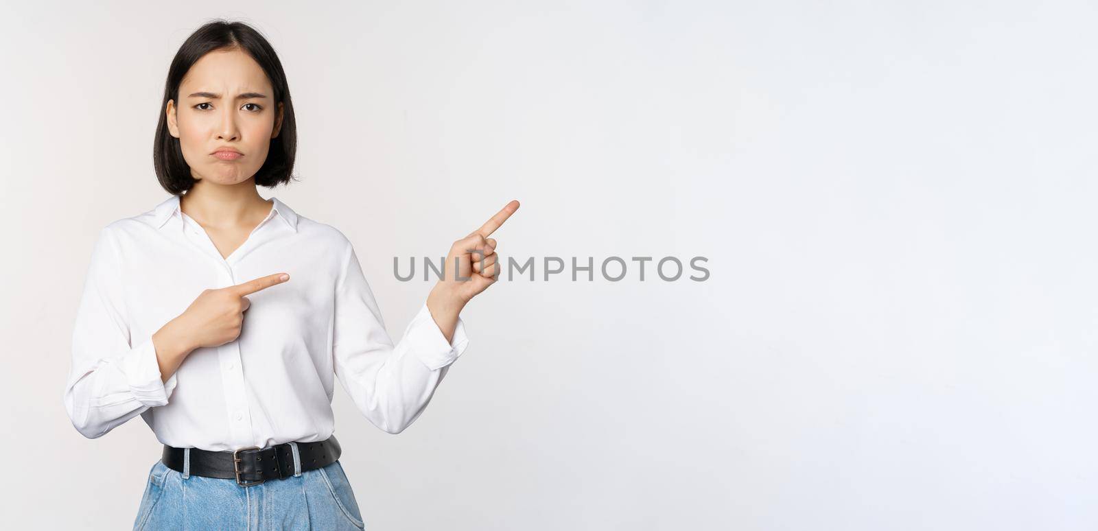 Sad and gloomy asian woman looking disappointed, complaining at banner or advertisement, pointing fingers right at promo and frowning upset, white background.