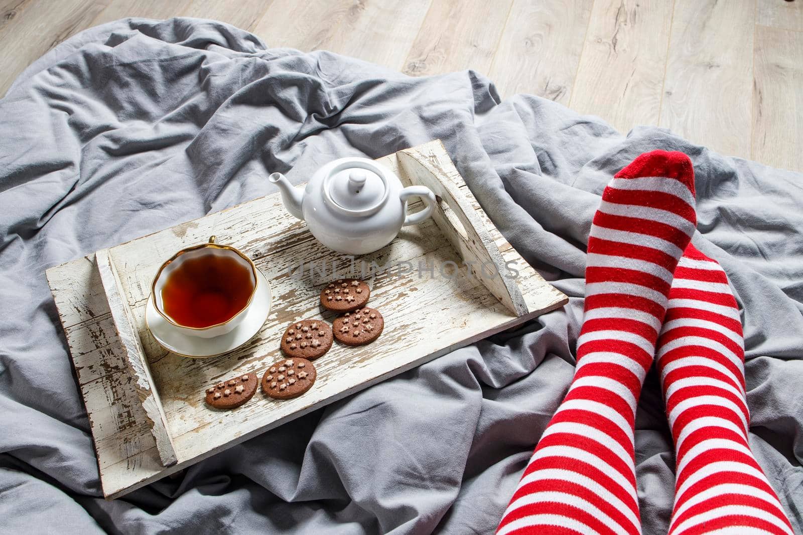 Fragment of legs in red striped socks. interior and home coziness concept. Top view. A cup of tea, a teapot with herbal tea, sugar bowl on a wooden white tray on the bed. Porcelain cup by elenarostunova