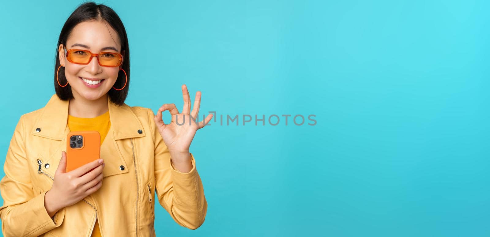 Smiling asian girl with smartphone, shwoing okay, ok sign in approval, standing over blue background. Copy space