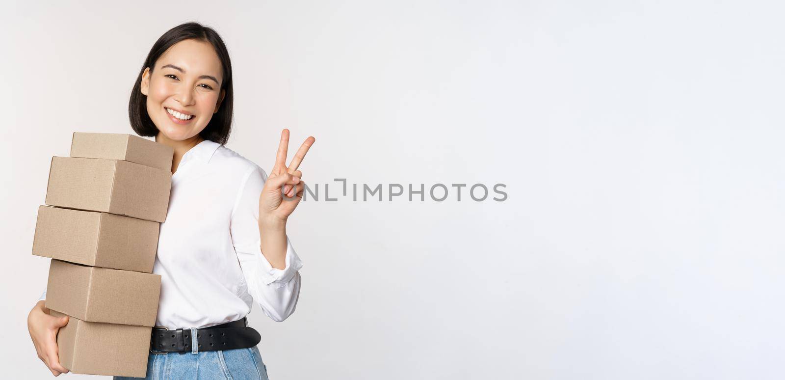 Smiling korean woman with boxes showing v-sign, peace gesture, standing over white background by Benzoix