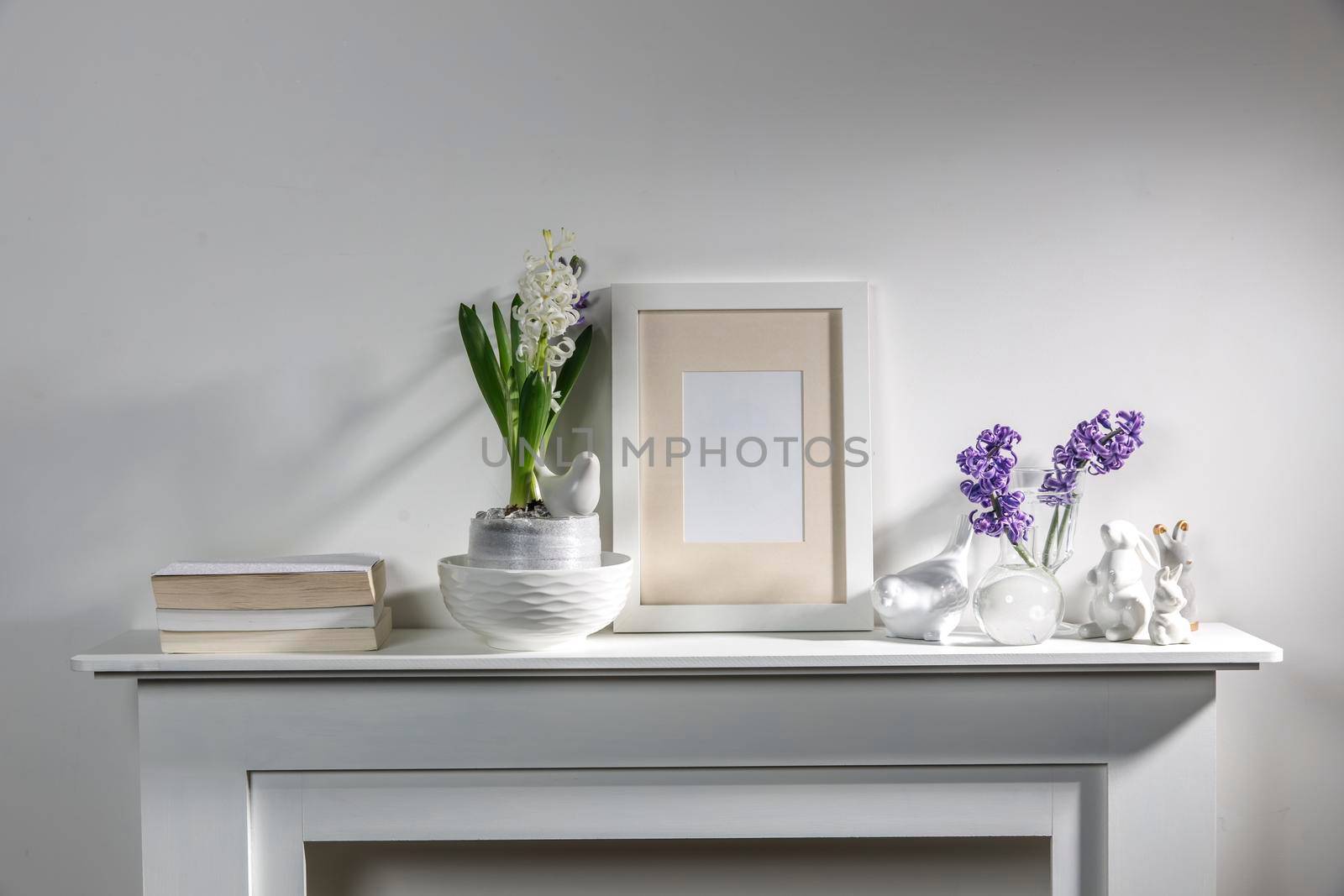 White hyacinth in a large porcelain bowl, books, figurines of hares and a bird, an empty photo frame are on the fireplace against the white wall. Layout.