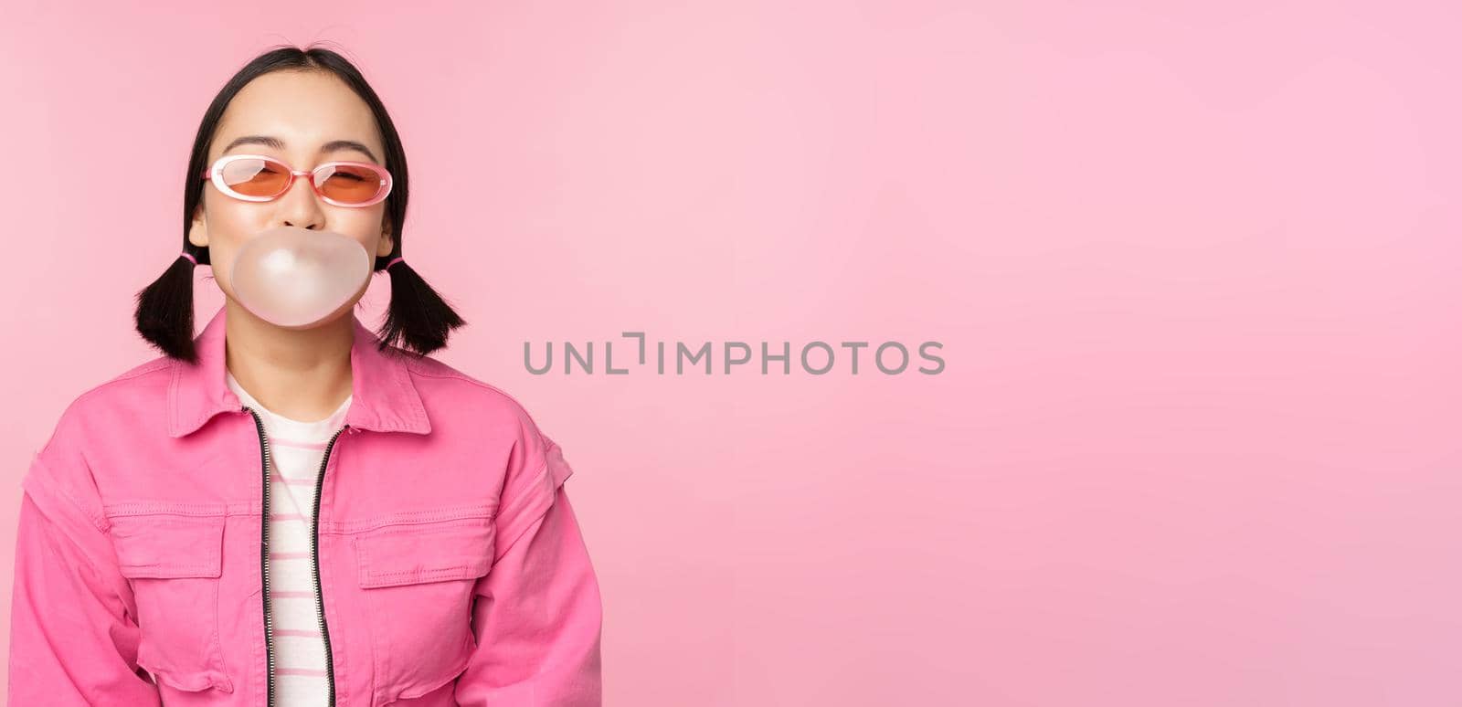 Stylish asian girl blowing bubblegum bubble, chewing gum, wearing sunglasses, posing against pink background. Copy space