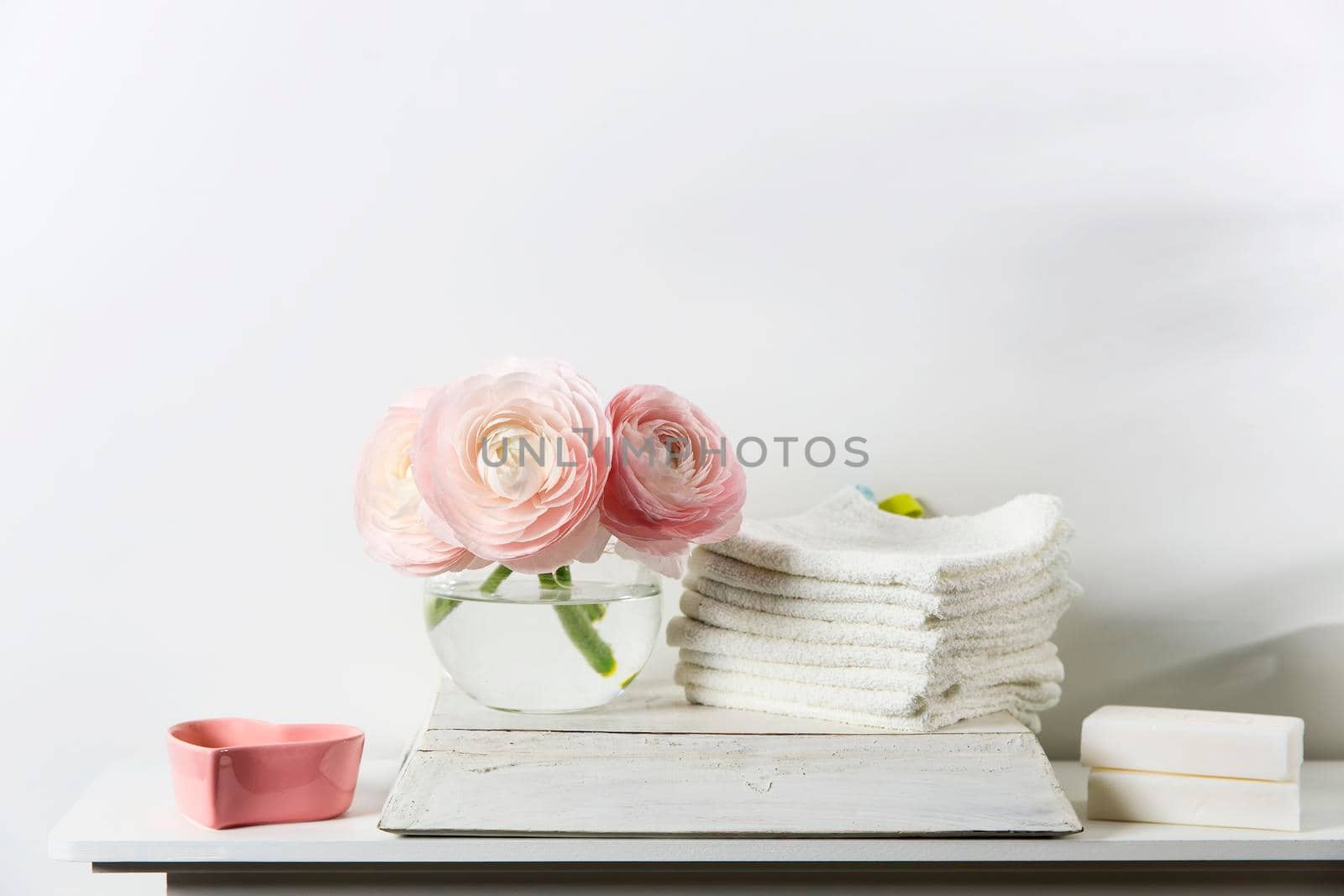 Three pale pink ranunculus in a transparent round vase and candles on the white windowsill. Rolled hand and face towel Copy space. Place for text by elenarostunova