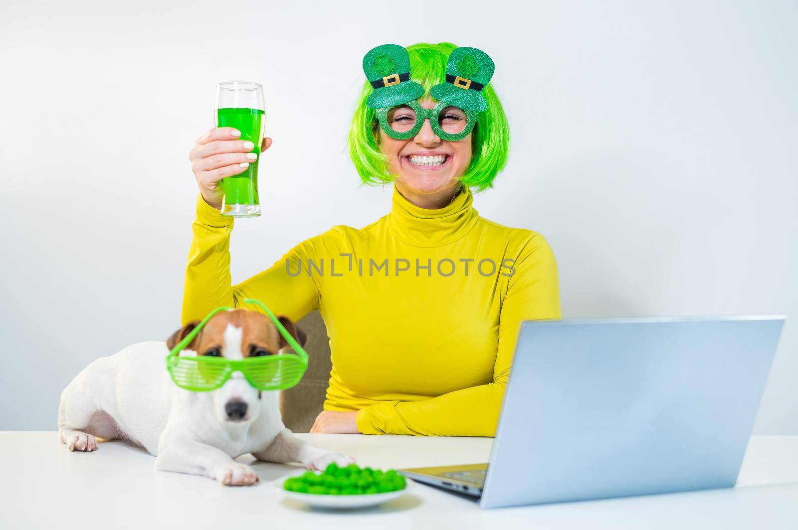 A young woman in a green wig and cheerful glasses drinks beer and bites glazed nuts. A girl sits with a dog at a table and celebrates st patrick's day online chatting with friends on a laptop. by mrwed54