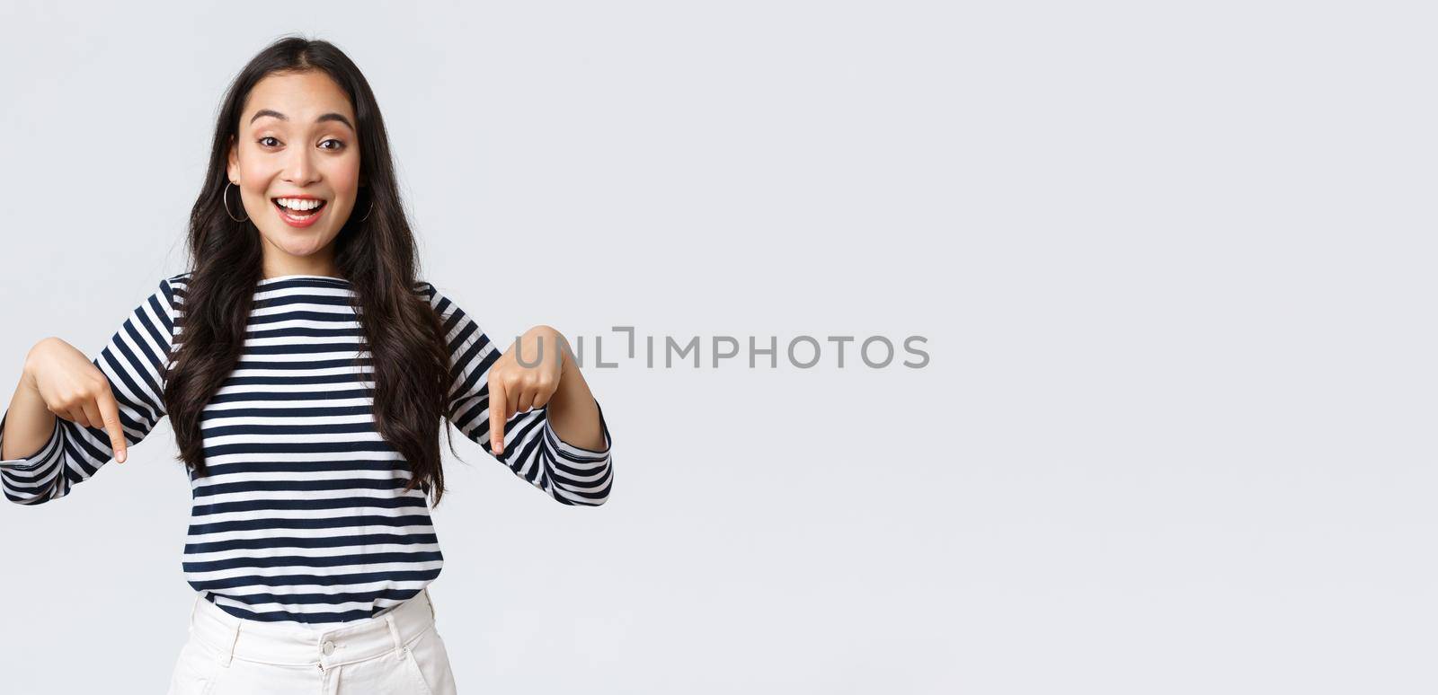 Lifestyle, beauty and fashion, people emotions concept. Excited tender charming asian girl with cute smile pointing fingers down, inviting customers to special event, white background.