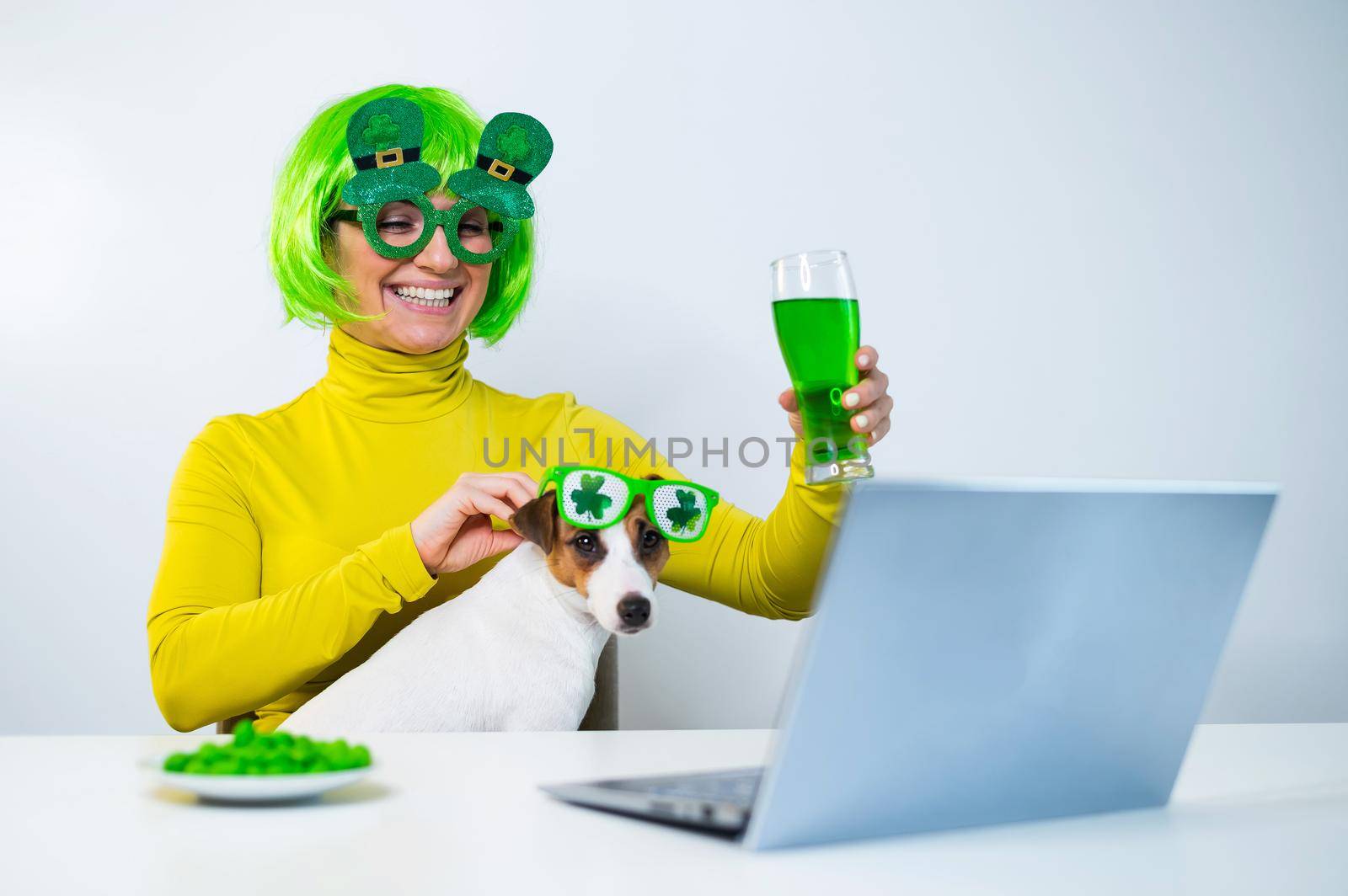 A young woman in a green wig and cheerful glasses drinks beer and bites glazed nuts. A girl sits with a dog at a table and celebrates st patrick's day online chatting with friends on a laptop. by mrwed54