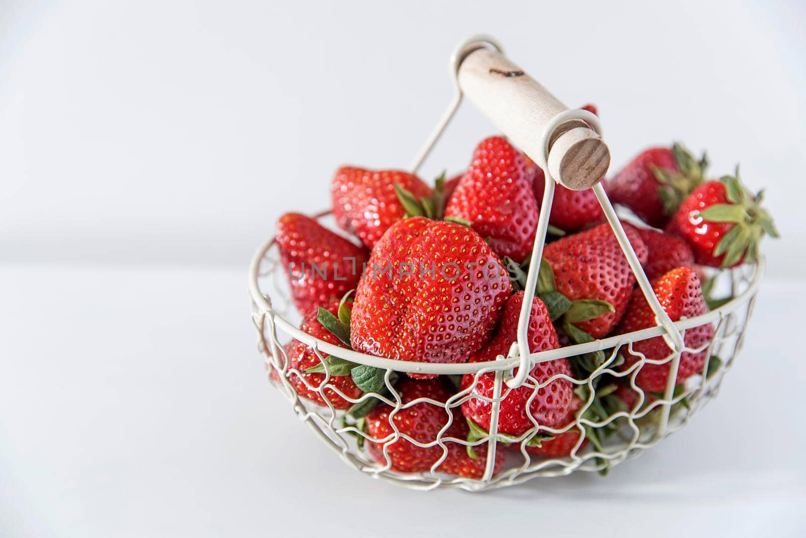 White metal basket with wooden handle with fresh strawberries is on the white table. Breakfast