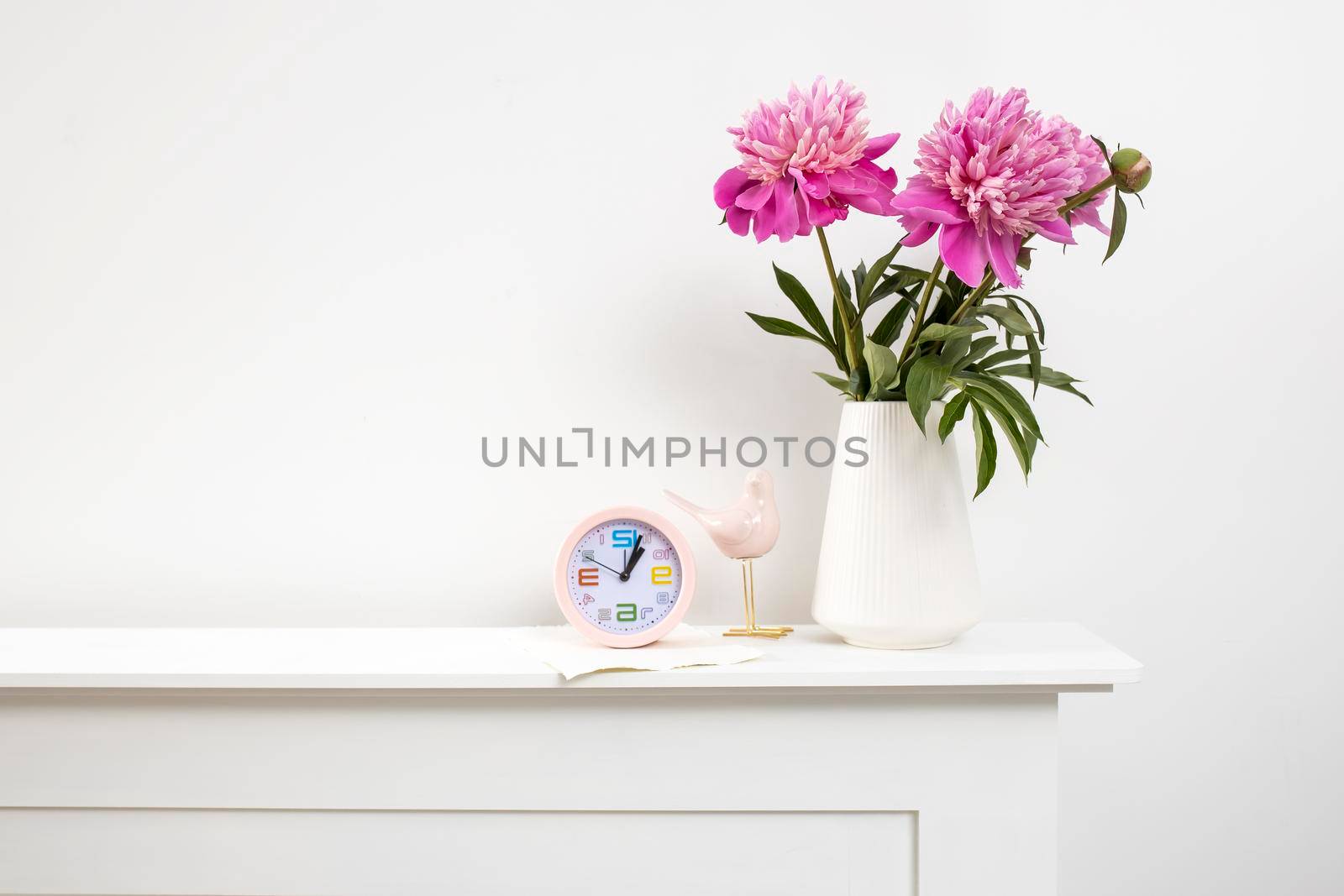 Red, pink peonies in a white vase on a table, clock, and against a white wall background by elenarostunova