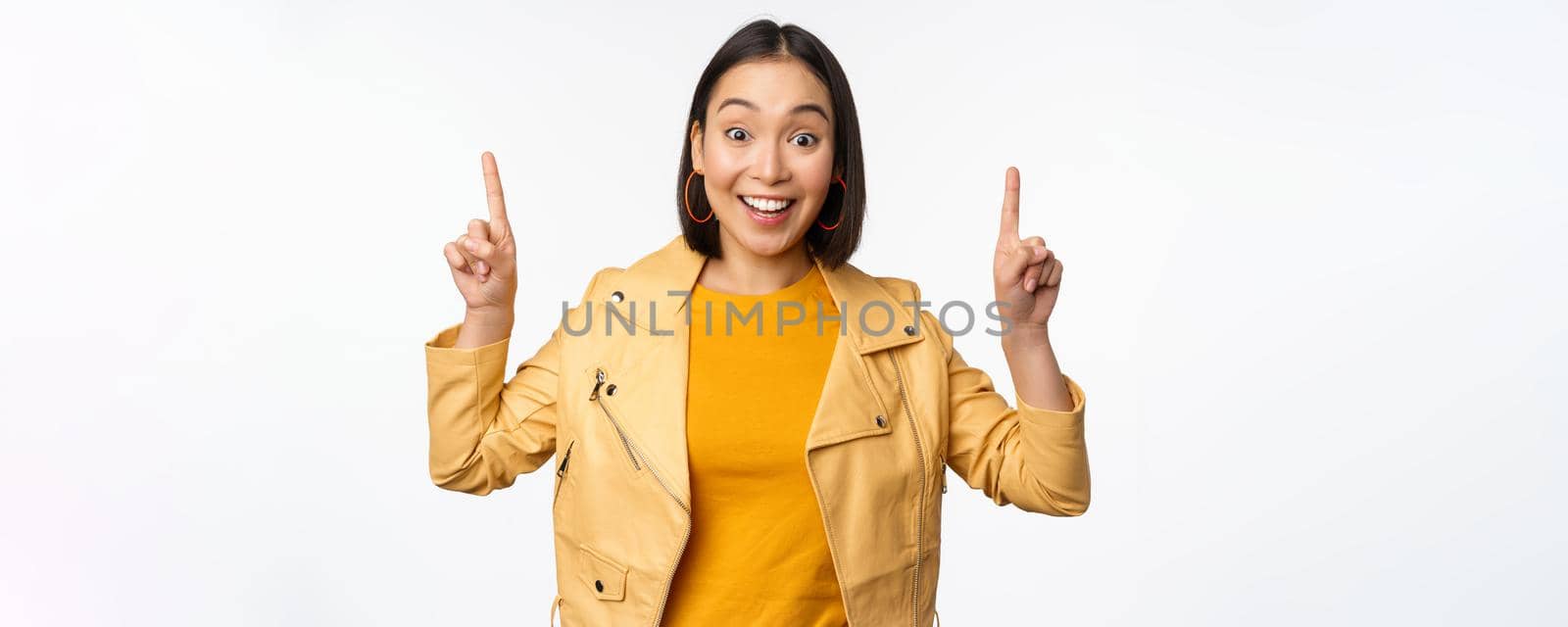 Enthusiastic asian girl pointing fingers up, showing advertisement on top, smiling happy, demonstrating promo offer or banner, standing over white background.