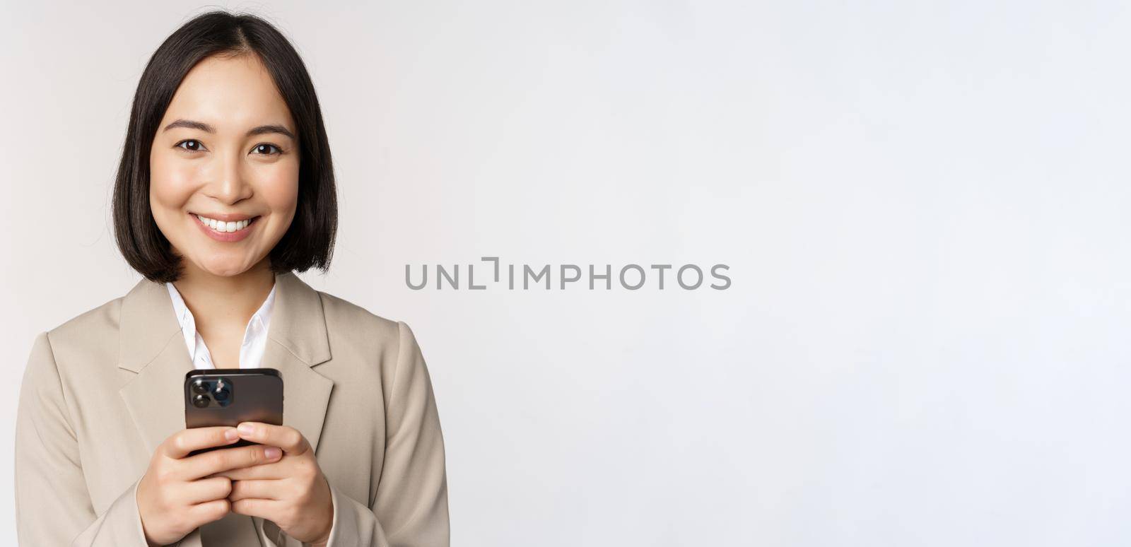 Image of asian businesswoman in suit, holding mobile phone, using smartphone app, smiling at camera, white background by Benzoix
