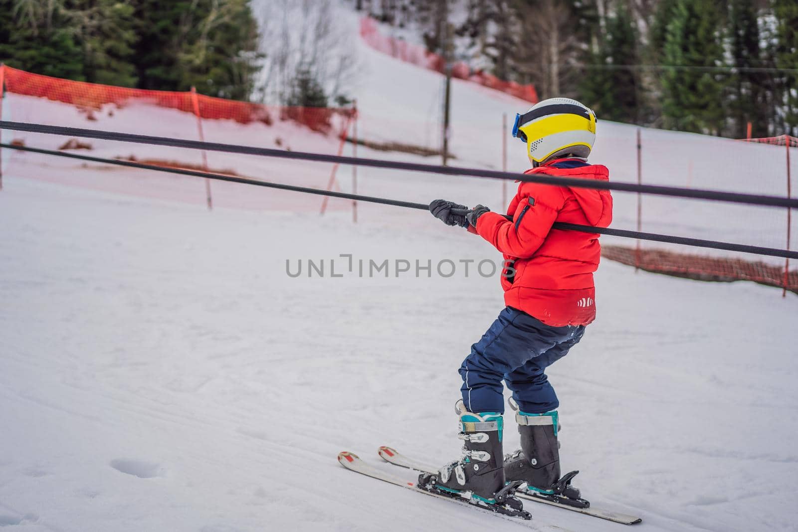 Boy uses a training lift. Child skiing in mountains. Active toddler kid with safety helmet, goggles and poles. Ski race for young children. Winter sport for family. Kids ski lesson in alpine school. Little skier racing in snow.