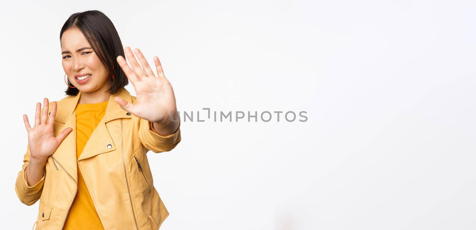 Stop, get away from me, back off concept. Young disgusted asian woman being blinded by smth, stretching out hands in defense, blocking light, standing over white background.