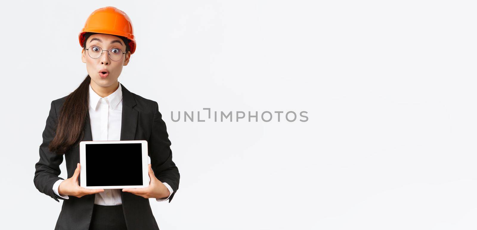 Impressed asian female construction engineer, businesswoman in safety helmet and suit showing chart, introduce digram at digial tablet screen, standing amazed over white background by Benzoix