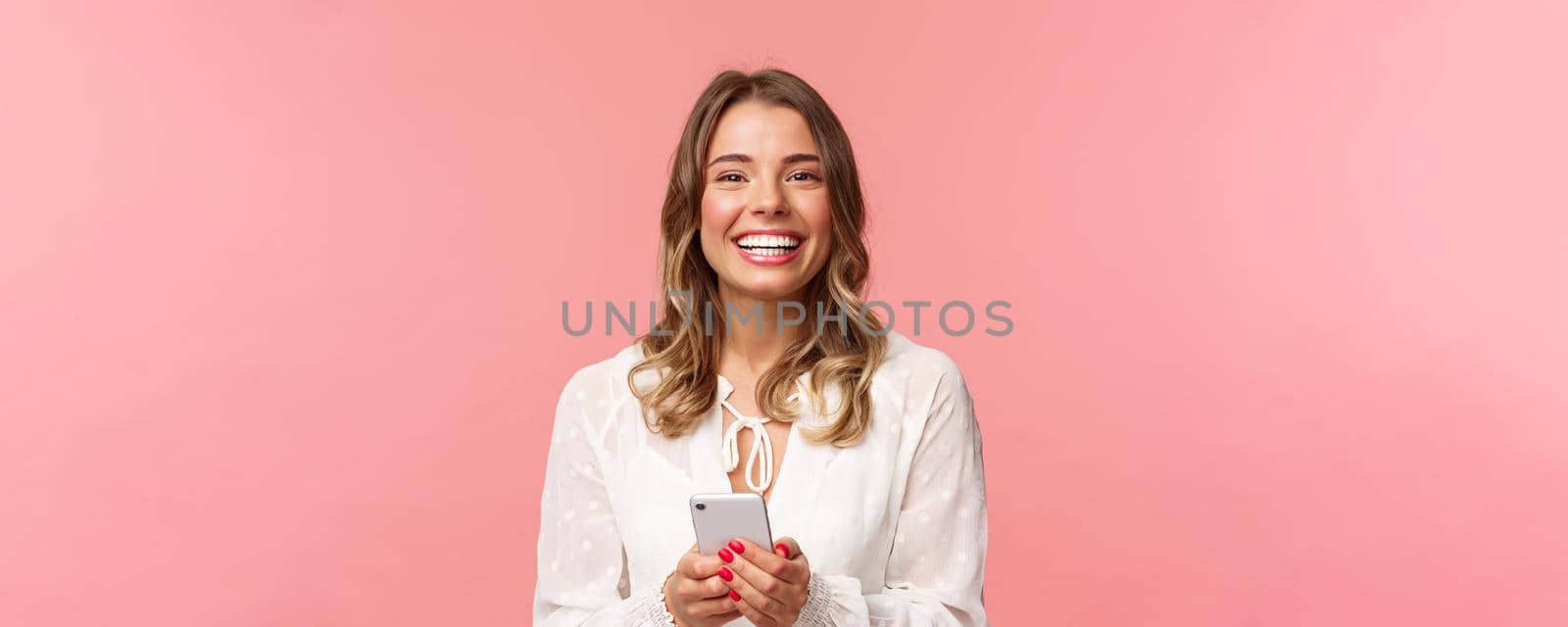 Close-up portrait of charming, lovely blond girl using mobile phone, texting friend, order in online store using smartphone application, laughing and smiling pleased, stand pink background by Benzoix
