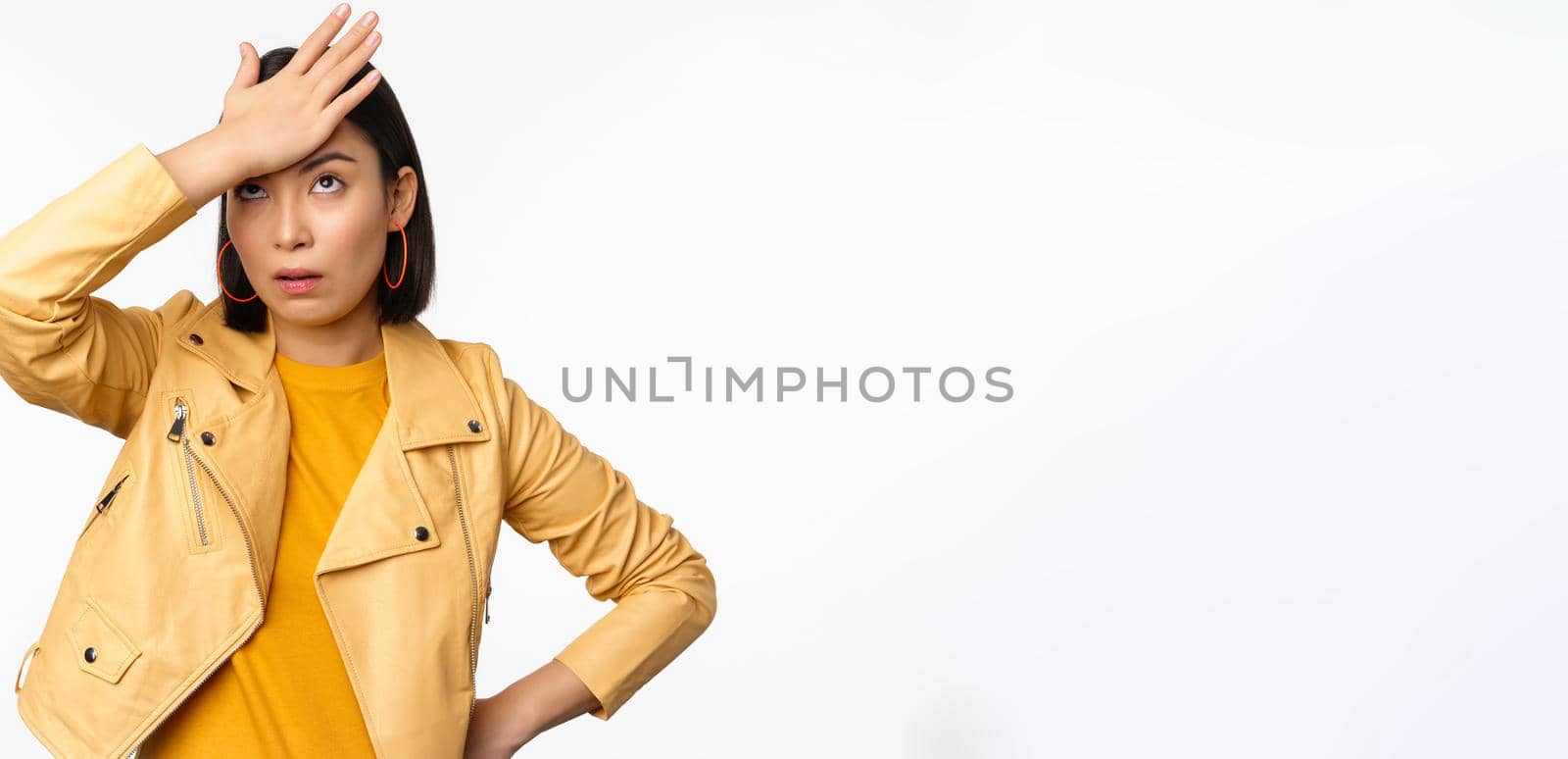 Annoyed and tired asian girl, slap forehead, facepalm and eyeroll with bothered face expression, standing in casual clothes over white studio background.
