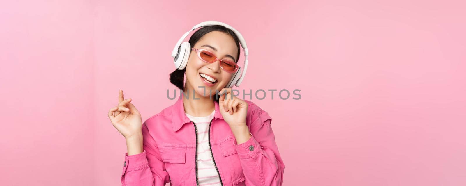 Dancing stylish asian girl listening music in headphones, posing against pink background.