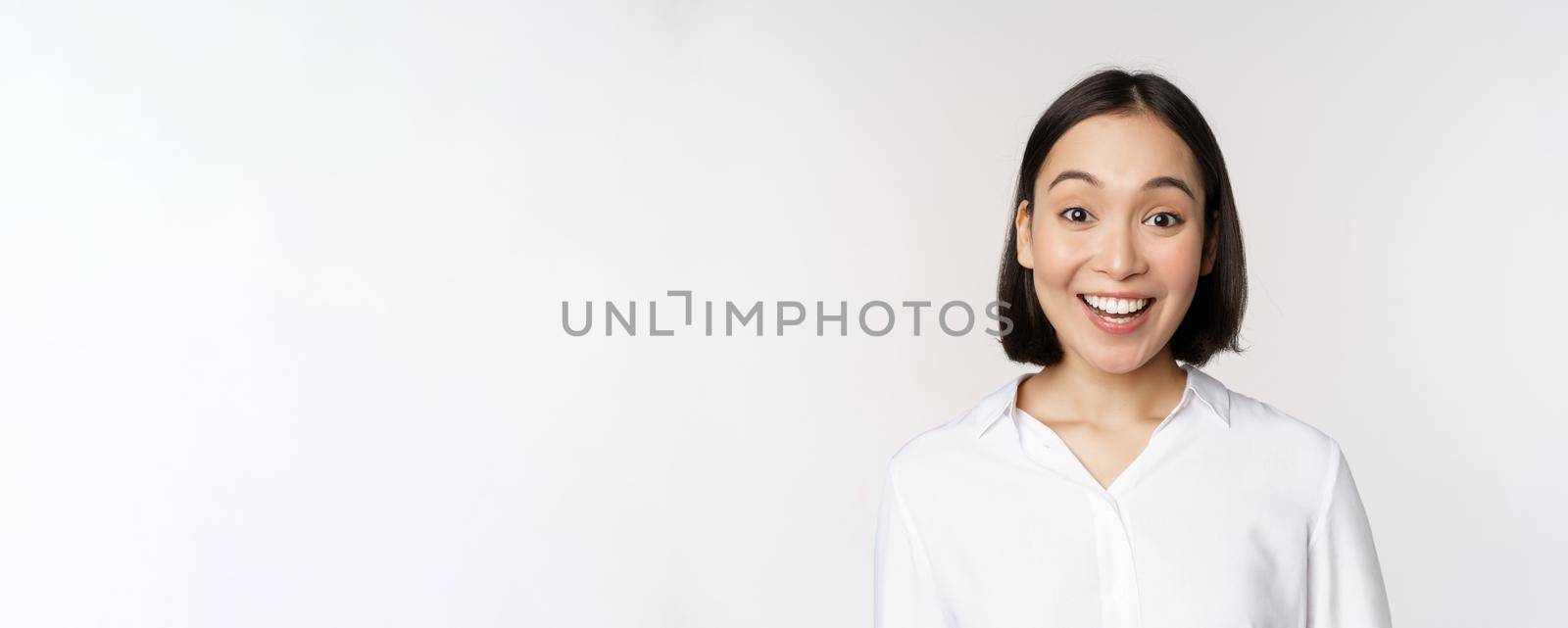 Close up portrait of young asian female model looking amazed at camera, smiling white teeth, standing against white background.
