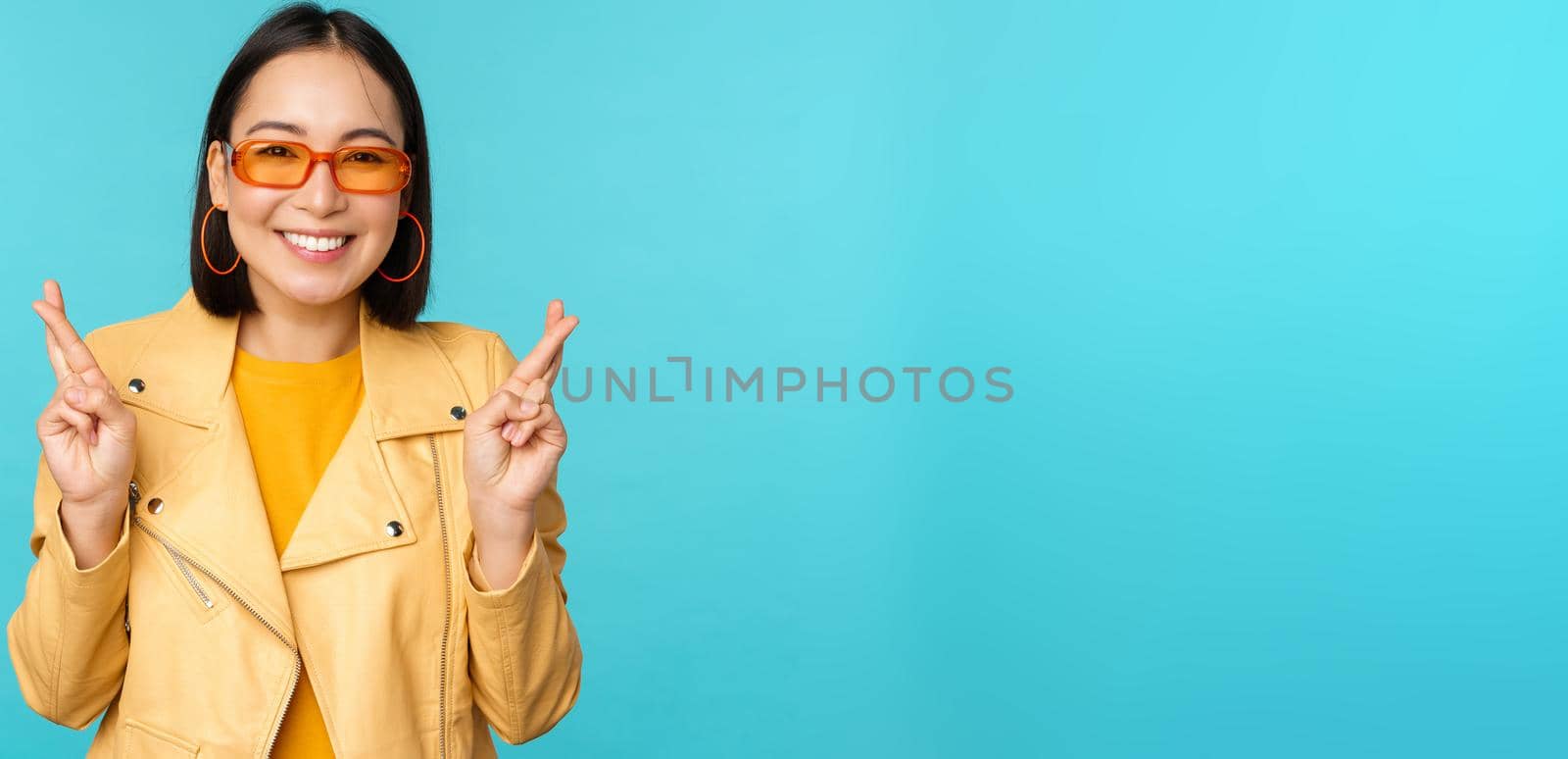 Smiling beautiful asian woman wishing, cross fingers for good luck and looking hopeful, standing over blue background. Copy space