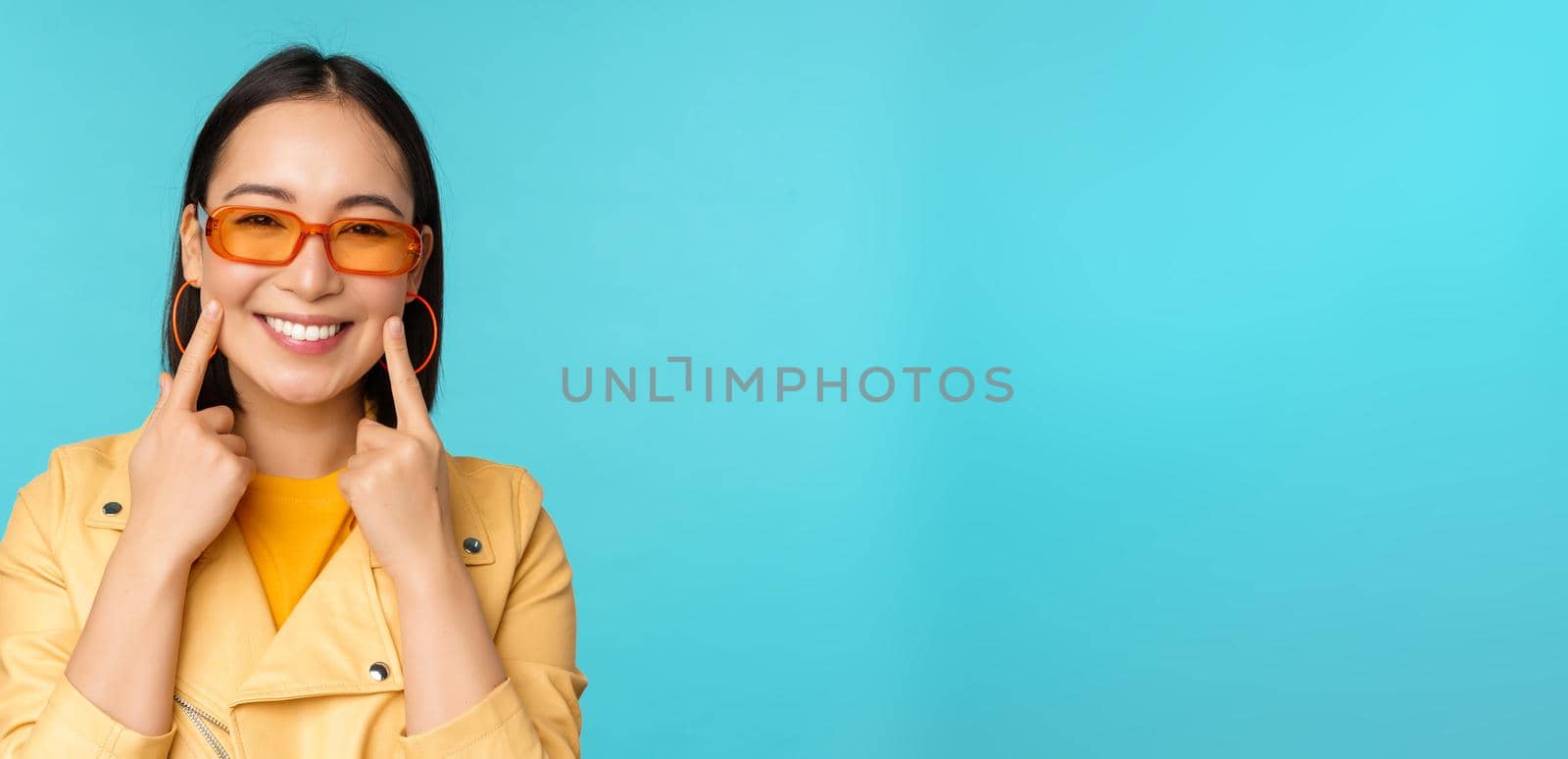 Close up portrait of asian young woman in sunglasses, smiling and looking romantic, standing happy over blue background. Copy space