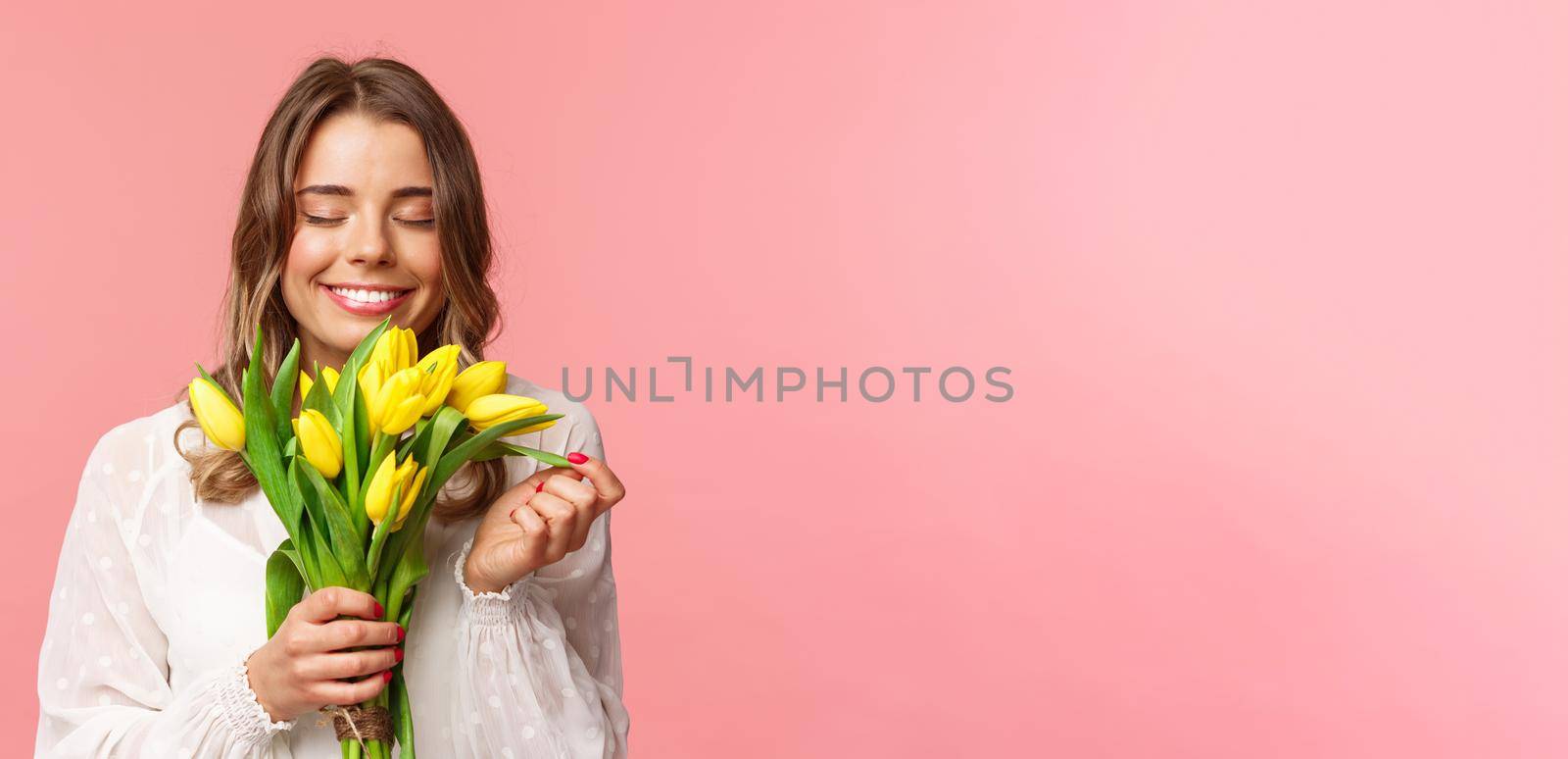 Spring, happiness and celebration concept. Close-up portrait of lovely, romantic blond girl sniffing smell of beautiful yellow tulips, close eyes and smiling happy, standing pink background.