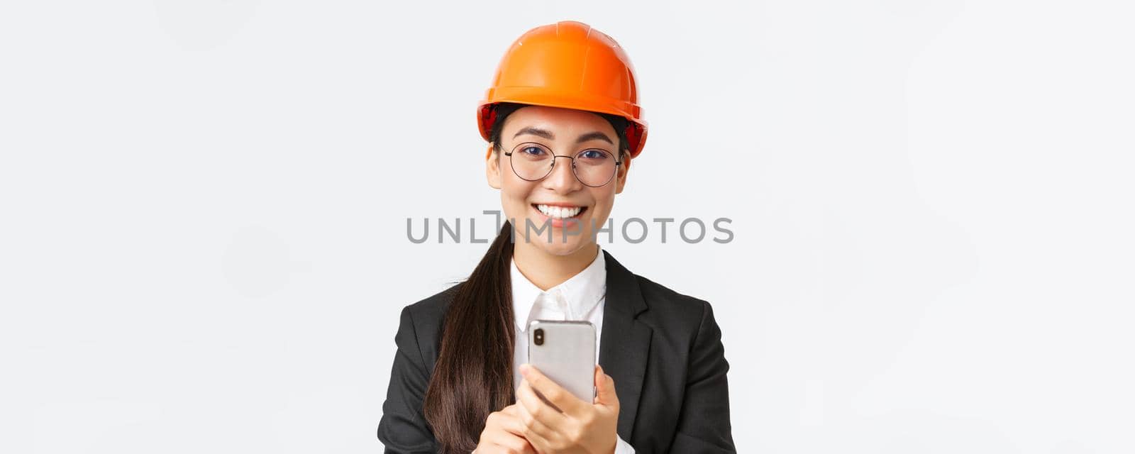 Close-up of successful female chief engineer, construction architect in safety helmet and business suit, glasses, looking at camera satisfied, smiling pleased as using mobile phone.