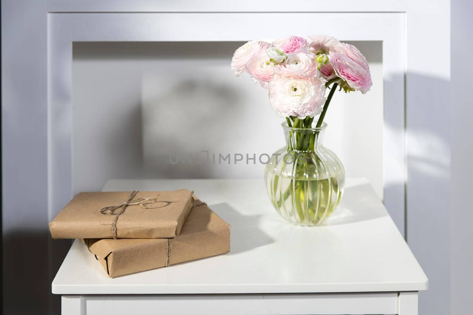 A bouquet of pink Persian buttercups in a glass vase and two wrapped gifts on a white table in front of a fake white fireplace