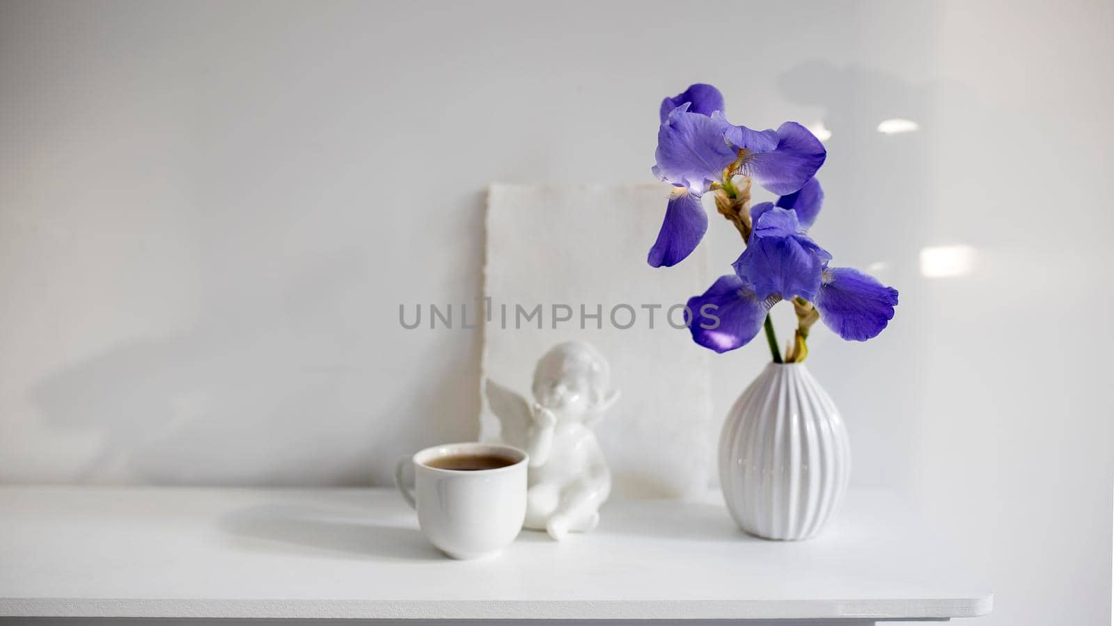 Blue iris in a corrugated vase with a piece of craft paper on a white table. Figurine of the angel. Cup of tea. Place for text