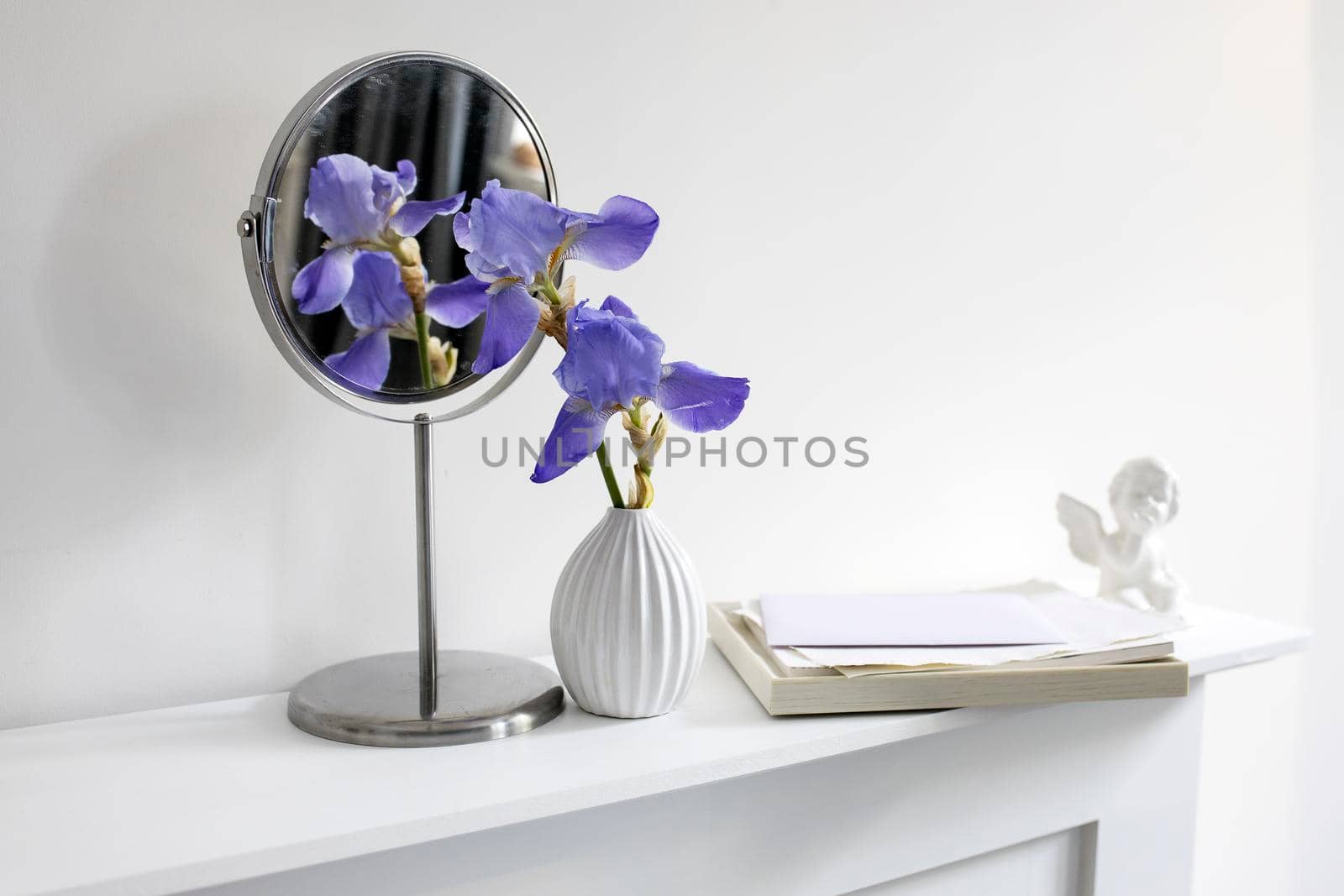 A branch of blue iris in a white fluted vase next to a round mirror, a figurine of an angel and a pile of white papers on a table against a white wall by elenarostunova