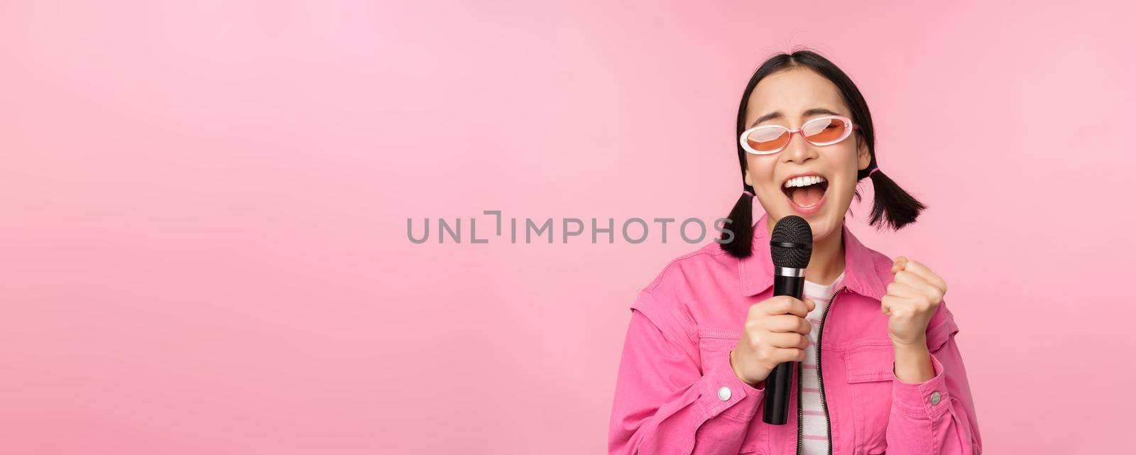 Happy beautiful asian girl singing with mic, using microphone, enjoying karaoke, posing against pink studio background.