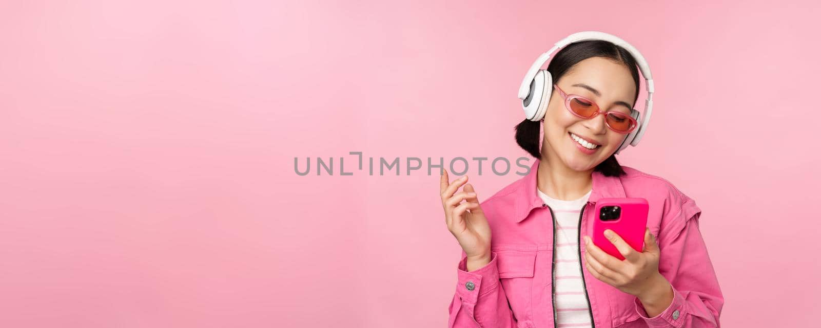 Stylish asian girl dancing with smartphone, listening music in headphones on mobile phone app, smiling and laughing, posing against pink background.
