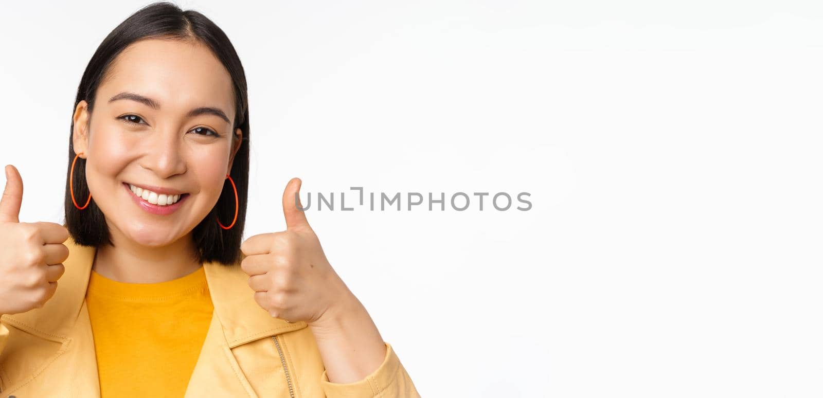 Smiling asian girl showing thumbs up, looking pleased, approve smth, standing over white background. Copy space