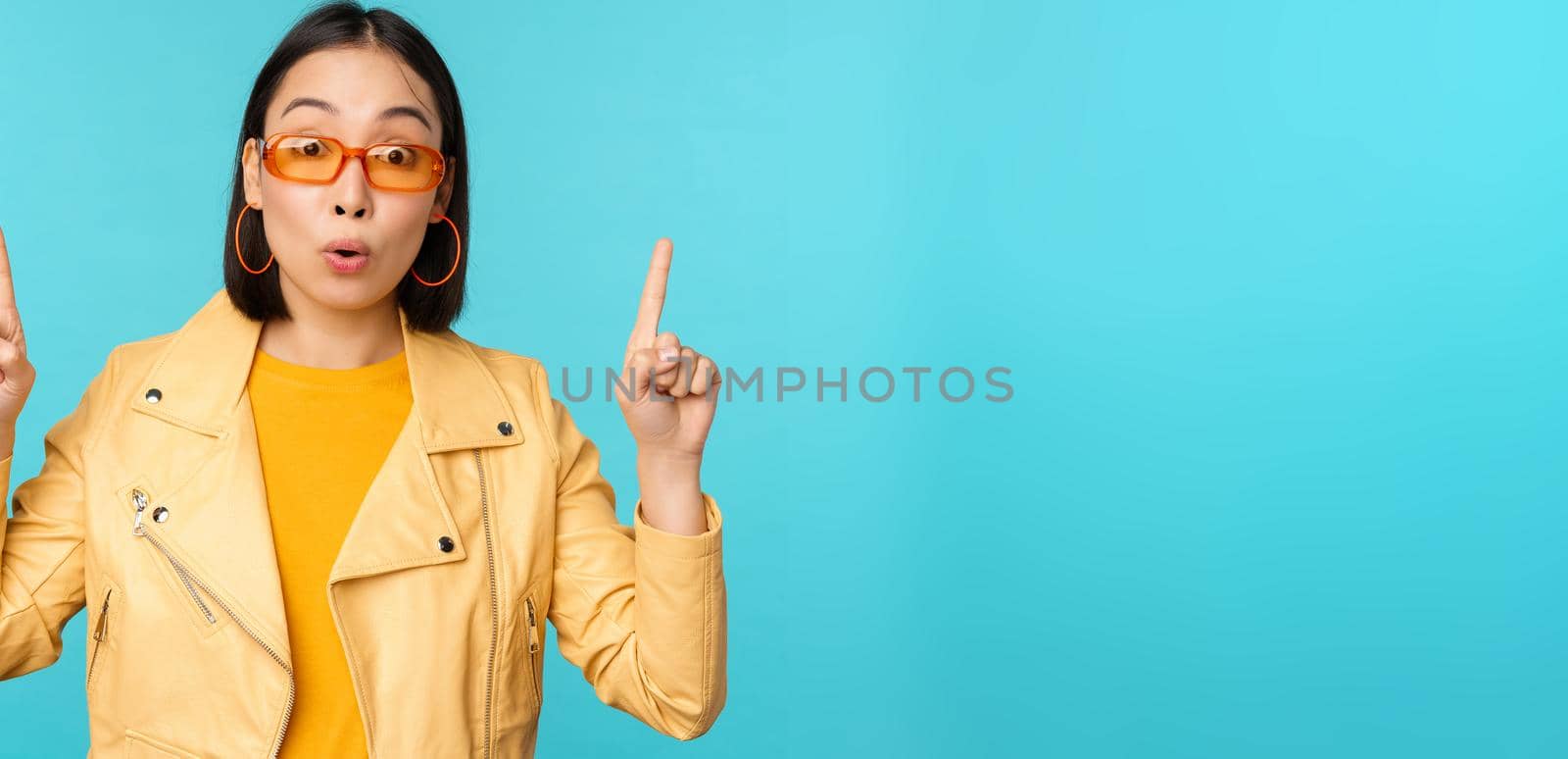 Enthusiastic asian girl in sunglasses, points fingers up, shows banner or logo on top, stands over blue background. Copy space