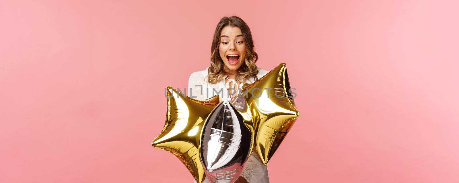 Holidays, celebration and women concept. Portrait of happy lovely young woman in white dress, gasping from amazement and joy, holding birthday star-shaped balloons, pink background.