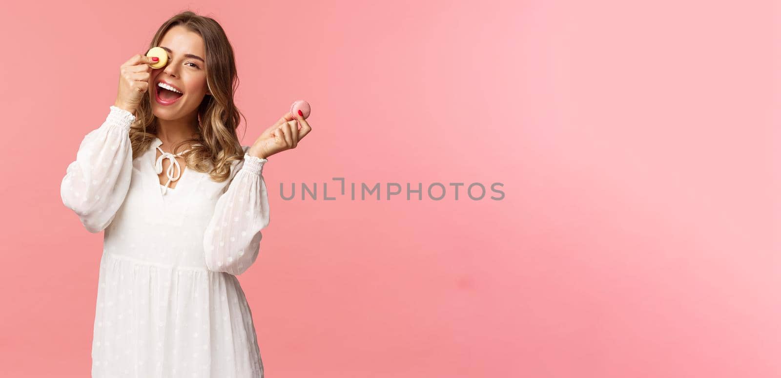 Holidays, spring and party concept. Portrait of carefree, tender and feminine girl in white dress, holding macarons dessert over eye, feeling cheerful, eating sweets, tasty desserts, pink background.