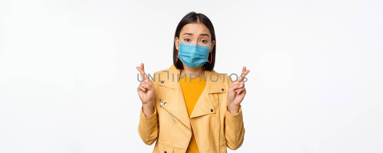 Hopeful asian girl in medical face mask, cross fingers, making wish, hoping, praying for smth, standing with anticipation over white studio background.