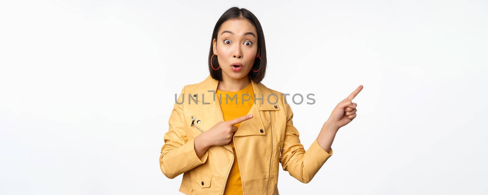 Enthusiastic asian girl pointing fingers right, showing advertisement and smiling, inviting to the store, standing over white background.