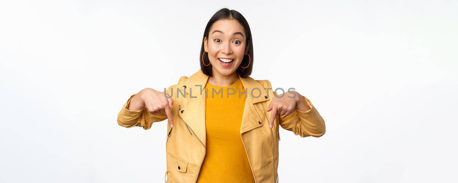 Portrait of happy smiling asian girl, pointing fingers down and showing logo, demonstrating banner, standing in yellow jacket against white background. Copy space