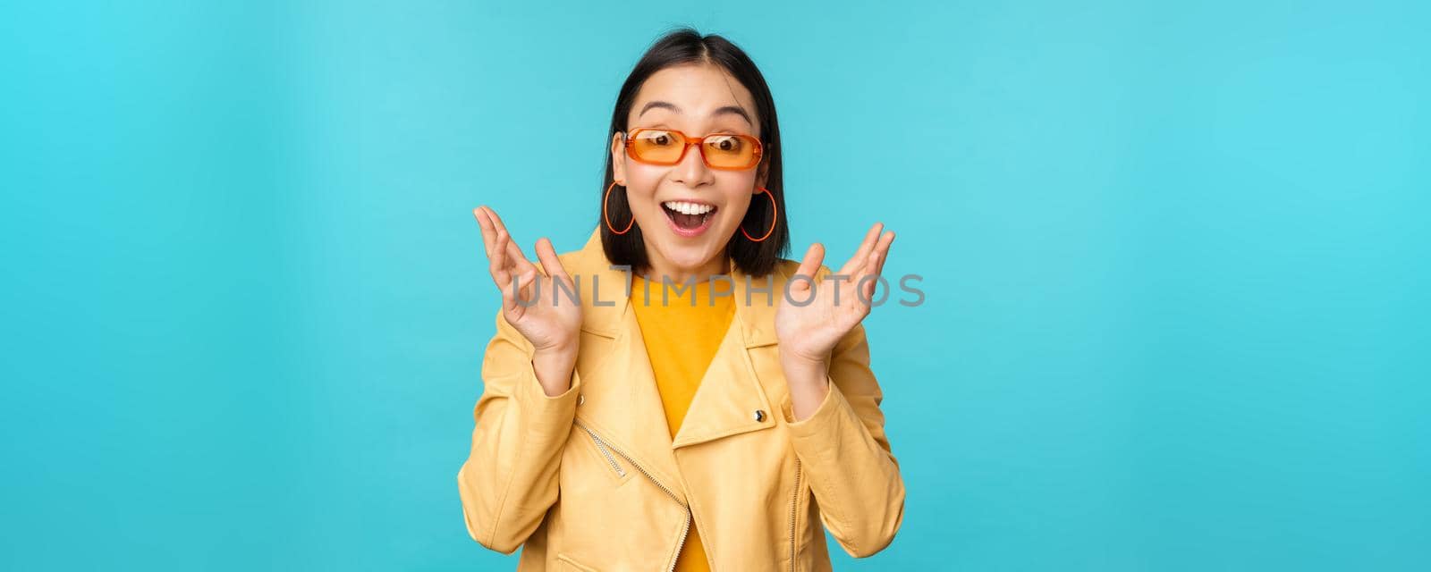 Image of enthusiastic young asian woman celebrating, triumphing, looking surprised and happy, clapping hands satisfied, standing over blue background.