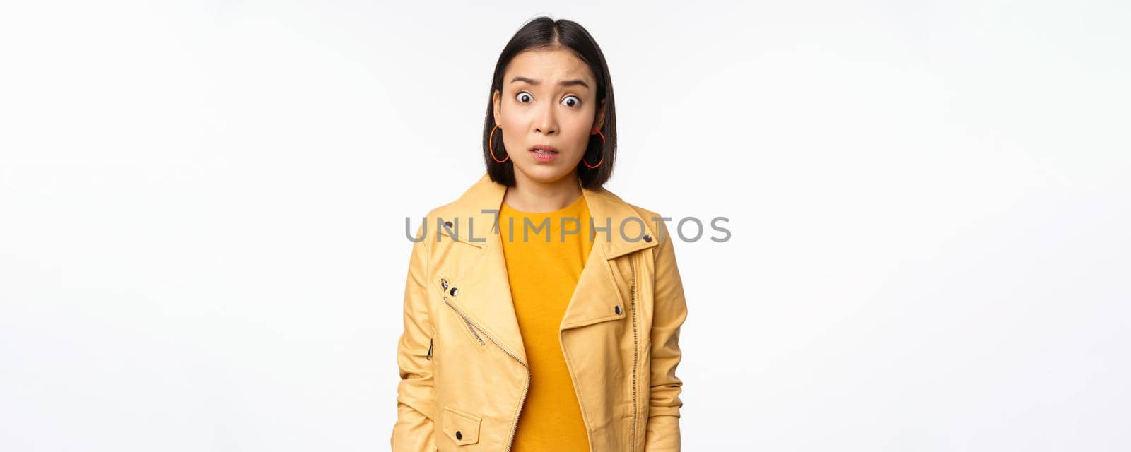 Image of shocked, confused asian woman, staring frustrated and speechless at camera, raising eyebrows and looking concerned, standing over white background.