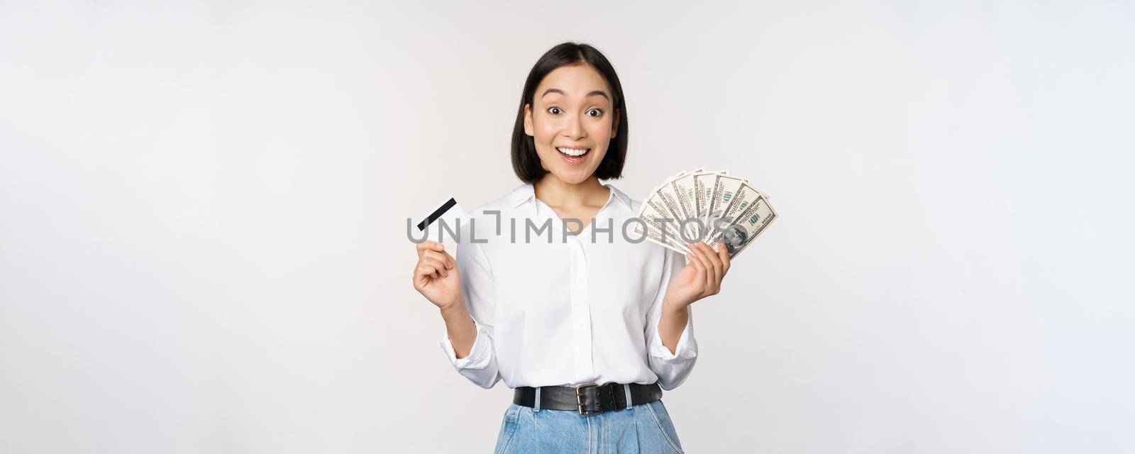 Portrait of enthusiastic asian woman holding money in cash and credit card, smiling amazed at camera, white background.