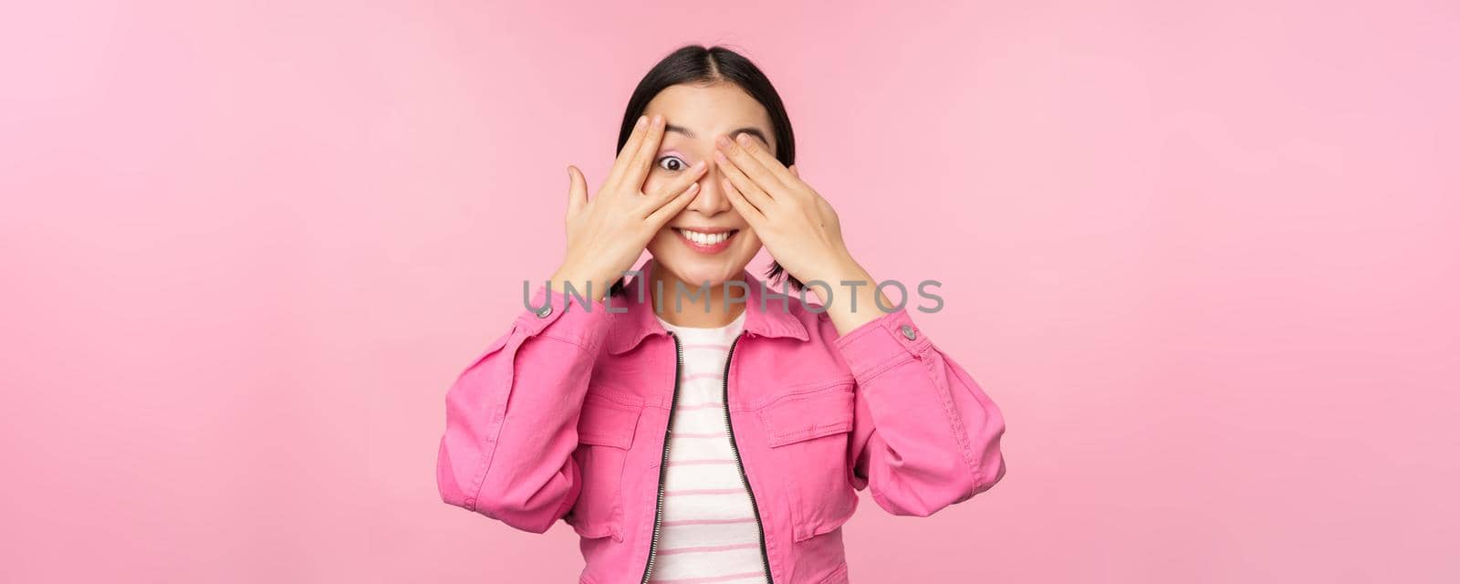 Portrait of asian girl peeks with excitement through fingers, covers eyes, seeing surprise, standing over pink background. Copy space