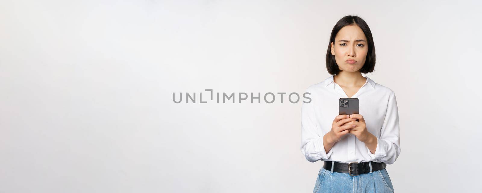 Asian woman holding smartphone and looking with doubt, disappointed with mobile phone app, standing agaist white background. Copy space