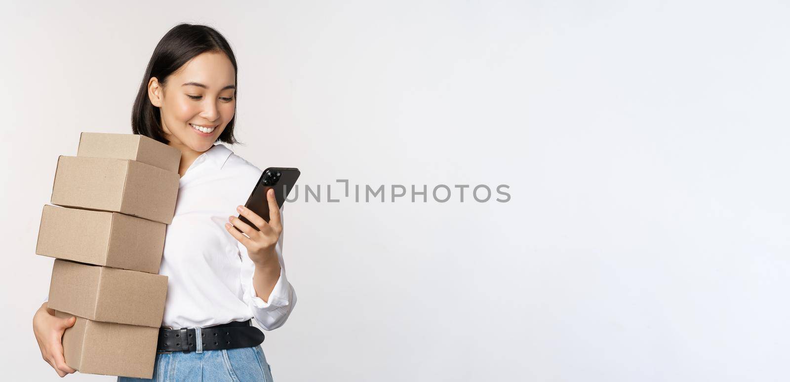 Image of young asian woman holding boxes, customer orders and looking at mobile phone, standing over white background.