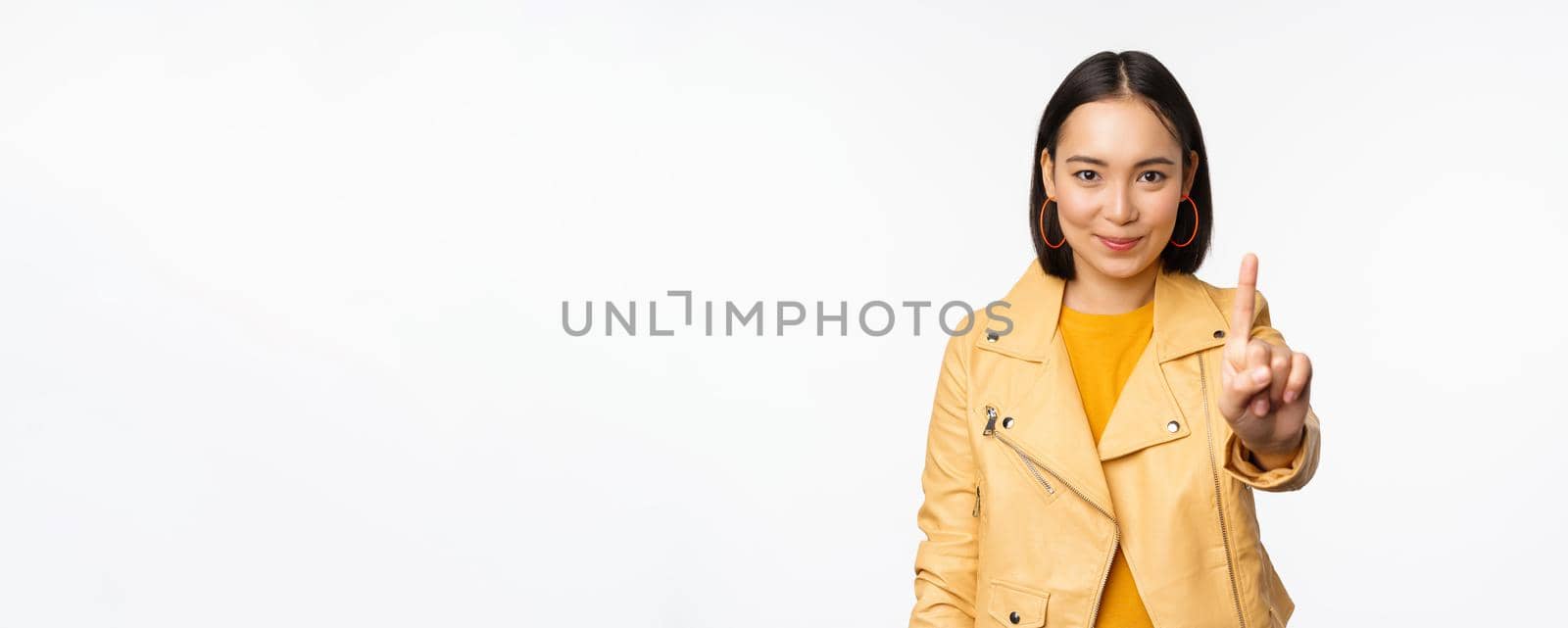 Rule number one. Confident smiling asian woman showing index finger, stop, prohibit gesture, disapprove smth, standing over white background.