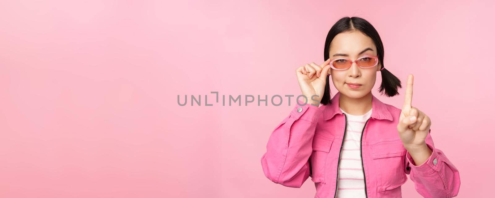 Image of asian girl with serious face, showing prohibit, scolding gesture, shaking finger pointing up, express disapproval, standing over pink background.