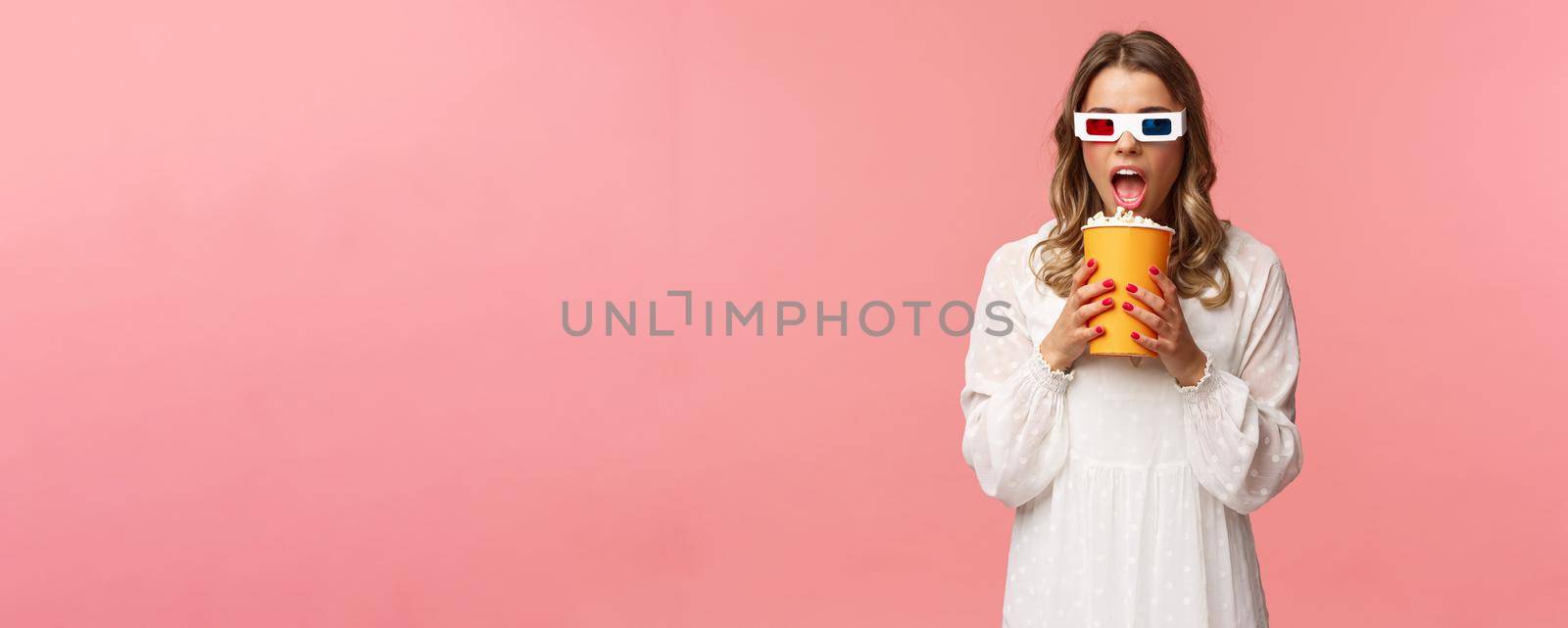 Leisure, going-out and spring concept. Portrait of cute blond funny girl in cinema, eating popcorn from box without hands, looking aside suspicious, wearing 3d glasses, pink background.