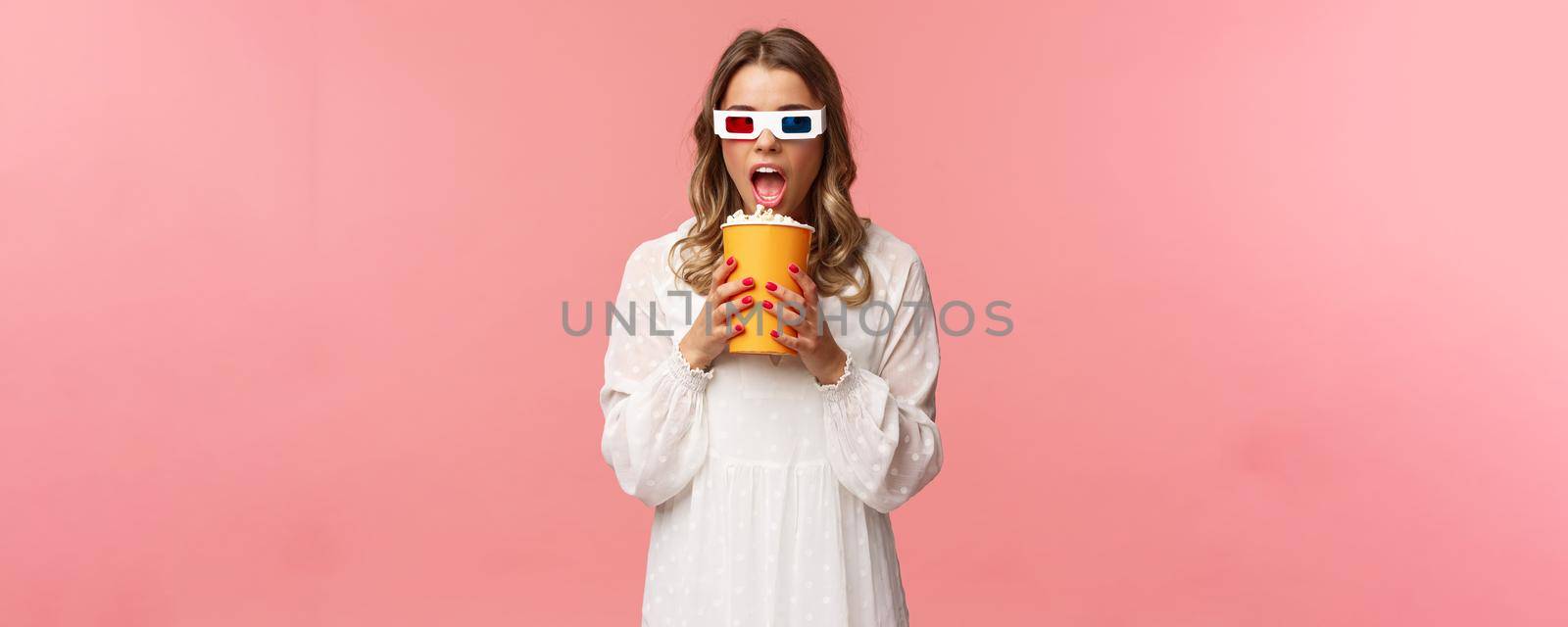 Leisure, going-out and spring concept. Portrait of cute blond funny girl in cinema, eating popcorn from box without hands, looking aside suspicious, wearing 3d glasses, pink background by Benzoix