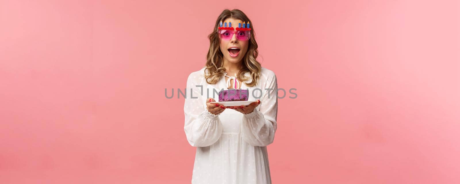Holidays, spring and party concept. Excited, happy attractive blond woman in white dress, wearing funny b-day glasses, holding birthday cake with lit candle, making wish smiling camera.