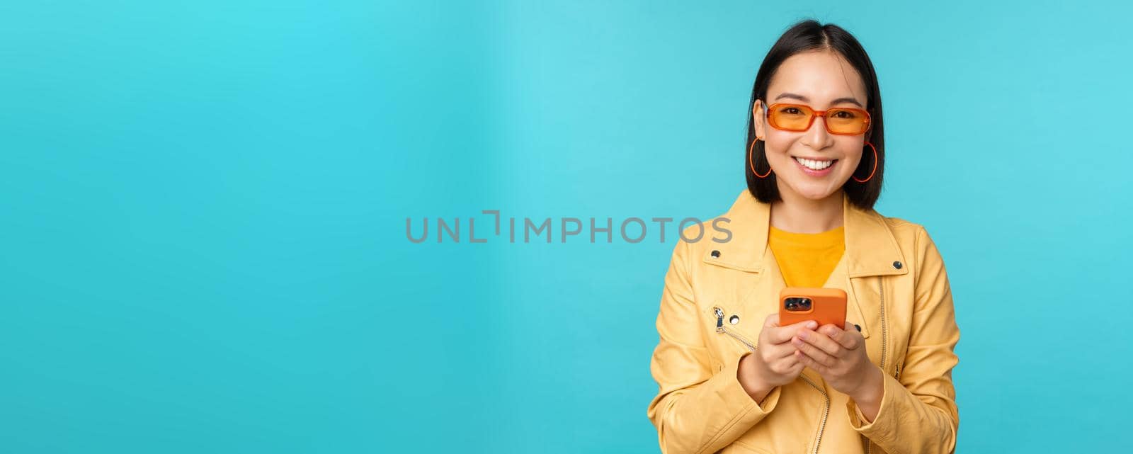 Smiling asian girl in sunglasses, using smartphone app, holding mobile phone, standing over blue background. Copy space