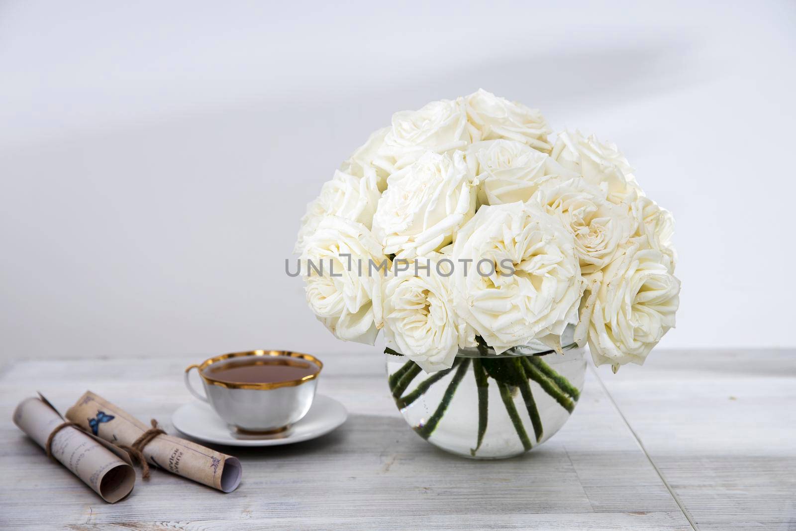 A bouquet of white roses in a round glass vase on a table with a cup of tea and a book. Copy space. by elenarostunova